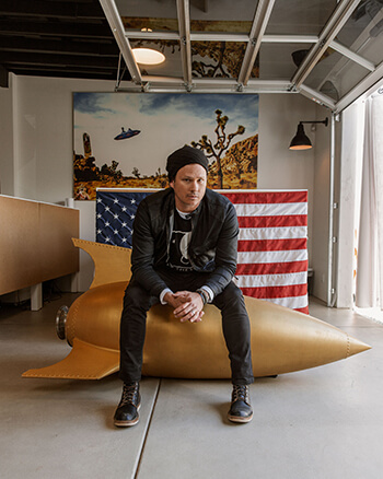 To the Stars founder Tom DeLonge sits atop a model rocket, behind him is an American flag draped over a dresser and a painting of a UFO in the desert.