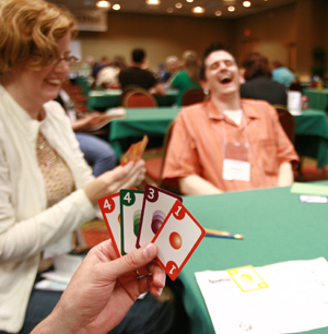 People playing a card game, a hand in the foreground holding cards while a man in the background laughs
