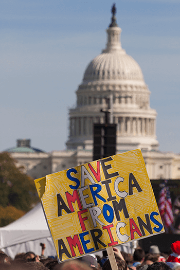 A person out of frame holds a sign that reads 'Save America from Americans' outside the U.S. Capitol.
